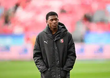Mukiele | AllezPaillade.com Young man wearing a black jacket with a team crest stands on a soccer pitch, hands in pockets.