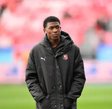Young man wearing a black jacket with a team crest stands on a soccer pitch, hands in pockets.