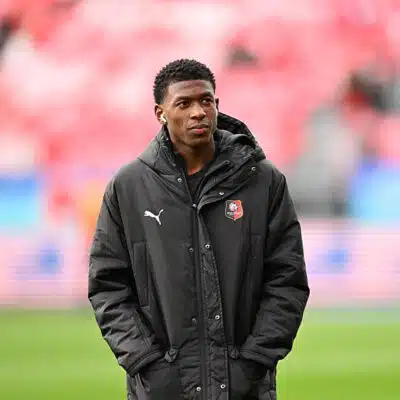 Young man wearing a black jacket with a team crest stands on a soccer pitch, hands in pockets.