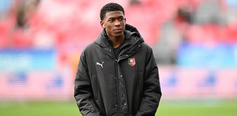 Mukiele | AllezPaillade.com Young man wearing a black jacket with a team crest stands on a soccer pitch, hands in pockets.