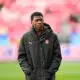 Young man wearing a black jacket with a team crest stands on a soccer pitch, hands in pockets.