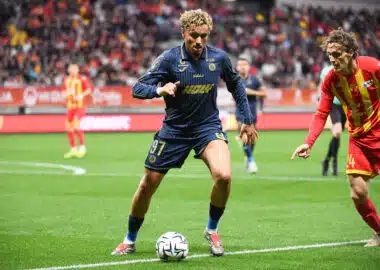 Soccer players chase the ball on a green pitch with a crowded stadium in the background.