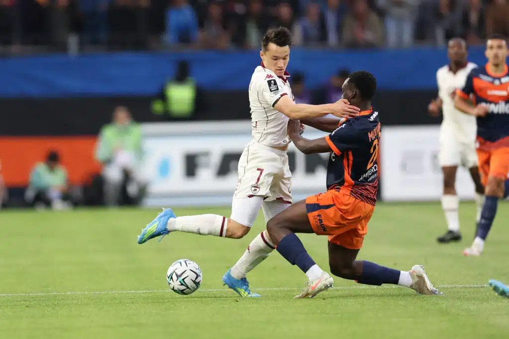 Mouanga Montpellier Herault SC v Clermont Foot 63 - Ligue 2 BKT 20252026 | AllezPaillade.com Two soccer players clash for the ball on a green pitch, one in a white kit and the other in orange-black, as spectators watch in the background.