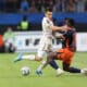 Two soccer players clash for the ball on a green pitch, one in a white kit and the other in orange-black, as spectators watch in the background.