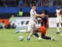 Two soccer players clash for the ball on a green pitch, one in a white kit and the other in orange-black, as spectators watch in the background.
