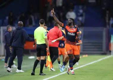 Soccer player in orange and navy kit lifts his right index finger while sprinting along the sideline, with teammates and officials nearby on the field.