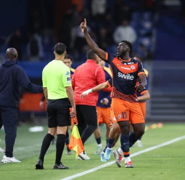 Soccer player in orange and navy kit lifts his right index finger while sprinting along the sideline, with teammates and officials nearby on the field.