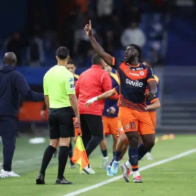 Soccer player in orange and navy kit lifts his right index finger while sprinting along the sideline, with teammates and officials nearby on the field.