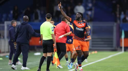 Soccer player in orange and navy kit lifts his right index finger while sprinting along the sideline, with teammates and officials nearby on the field.