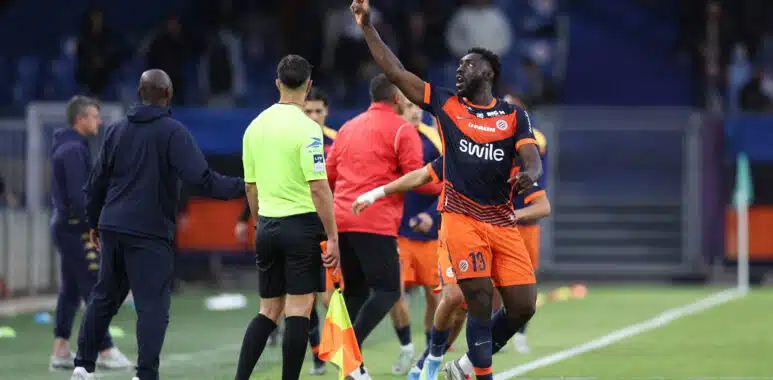 Alexandre Mendy Montpellier Herault SC v Clermont Foot 63 - Ligue 2 BKT 20252026 | AllezPaillade.com Soccer player in orange and navy kit lifts his right index finger while sprinting along the sideline, with teammates and officials nearby on the field.