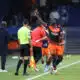 Soccer player in orange and navy kit lifts his right index finger while sprinting along the sideline, with teammates and officials nearby on the field.