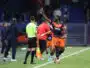 Soccer player in orange and navy kit lifts his right index finger while sprinting along the sideline, with teammates and officials nearby on the field.
