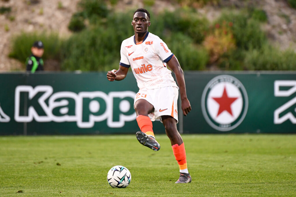 Mouanga Red Star FC v Montpellier Herault SC - Ligue 2 BKT 20252026 | AllezPaillade.com Soccer player in a white kit with orange accents dribbles a ball on a green field during a match, with a dark-haired player in the background.