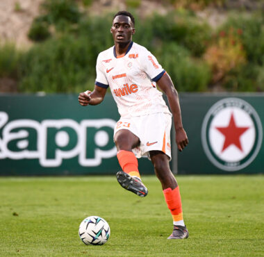 Soccer player in a white kit with orange accents dribbles a ball on a green field during a match, with a dark-haired player in the background.