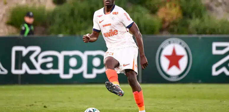 Mouanga Red Star FC v Montpellier Herault SC - Ligue 2 BKT 20252026 | AllezPaillade.com Soccer player in a white kit with orange accents dribbles a ball on a green field during a match, with a dark-haired player in the background.