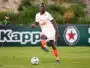 Soccer player in a white kit with orange accents dribbles a ball on a green field during a match, with a dark-haired player in the background.