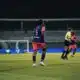 Soccer player wearing blue jersey number 5 stands on a green field during a night match, near teammates and a goalie in the background.