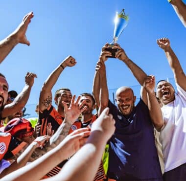 A diverse team of athletes in colorful jerseys cheer and lift a silver trophy high above their heads against a bright blue sky.