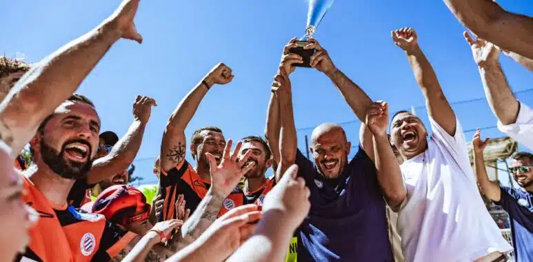 IMG_5173 | AllezPaillade.com A diverse team of athletes in colorful jerseys cheer and lift a silver trophy high above their heads against a bright blue sky.