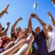 A diverse team of athletes in colorful jerseys cheer and lift a silver trophy high above their heads against a bright blue sky.
