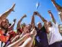 A diverse team of athletes in colorful jerseys cheer and lift a silver trophy high above their heads against a bright blue sky.