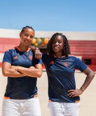 IMG_5196 | AllezPaillade.com Two women in navy athletic shirts with orange trim stand on a sunny outdoor court, arms around each other.