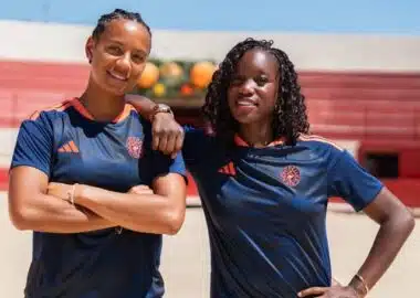 Two women in navy athletic shirts with orange trim stand on a sunny outdoor court, arms around each other.