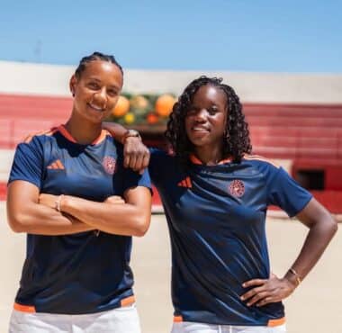 Two women in navy athletic shirts with orange trim stand on a sunny outdoor court, arms around each other.