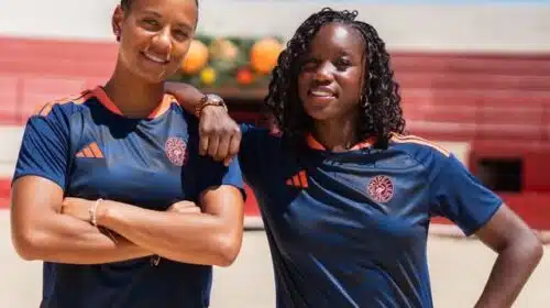 Two women in navy athletic shirts with orange trim stand on a sunny outdoor court, arms around each other.