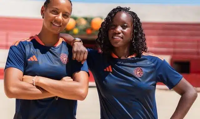 IMG_5196 | AllezPaillade.com Two women in navy athletic shirts with orange trim stand on a sunny outdoor court, arms around each other.