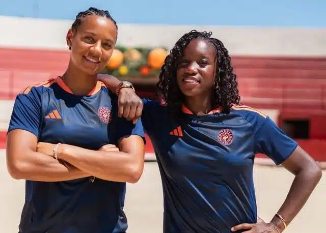 Two women in navy athletic shirts with orange trim stand on a sunny outdoor court, arms around each other.