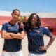 Two women in navy athletic shirts with orange trim stand on a sunny outdoor court, arms around each other.