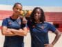 Two women in navy athletic shirts with orange trim stand on a sunny outdoor court, arms around each other.