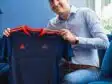 Smiling man in a light blue shirt holds up a dark blue sports jersey with orange trim indoors.