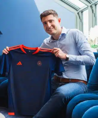 Smiling man in a light blue shirt holds up a dark blue sports jersey with orange trim indoors.