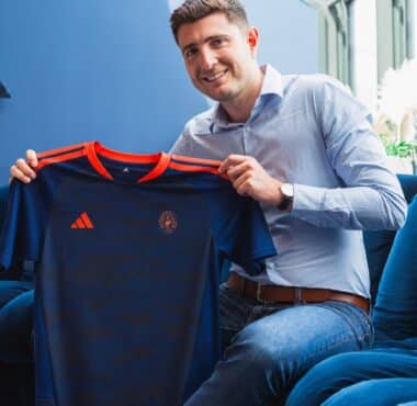 Smiling man in a light blue shirt holds up a dark blue sports jersey with orange trim indoors.