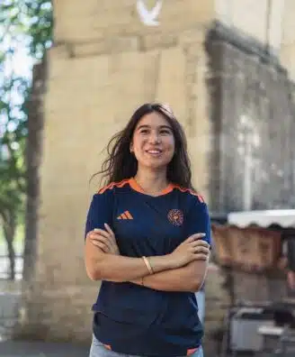 Smiling woman with arms crossed outdoors, wearing a navy athletic shirt with orange accents near a stone archway.