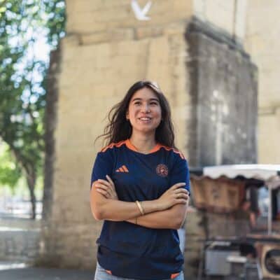 Smiling woman with arms crossed outdoors, wearing a navy athletic shirt with orange accents near a stone archway.