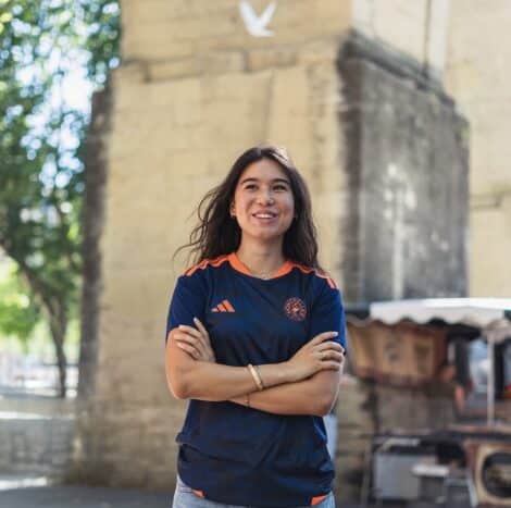 Smiling woman with arms crossed outdoors, wearing a navy athletic shirt with orange accents near a stone archway.