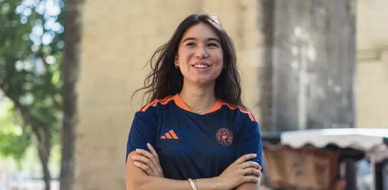 Screenshot | AllezPaillade.com Smiling woman with arms crossed outdoors, wearing a navy athletic shirt with orange accents near a stone archway.