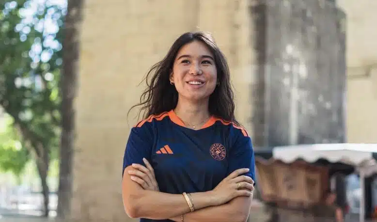 Screenshot | AllezPaillade.com Smiling woman with arms crossed outdoors, wearing a navy athletic shirt with orange accents near a stone archway.