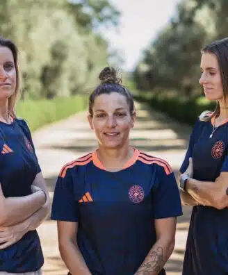 Screenshot | AllezPaillade.com Three women in matching navy blue sports jerseys with orange accents stand on a sunlit, tree-lined path, arms crossed and facing the camera.