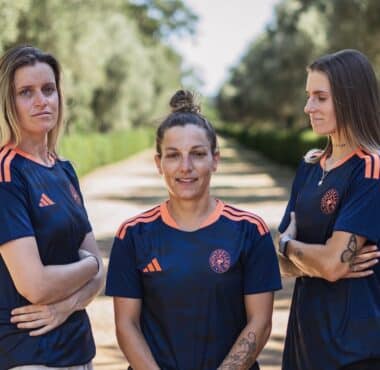 Three women in matching navy blue sports jerseys with orange accents stand on a sunlit, tree-lined path, arms crossed and facing the camera.