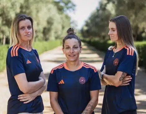 Three women in matching navy blue sports jerseys with orange accents stand on a sunlit, tree-lined path, arms crossed and facing the camera.