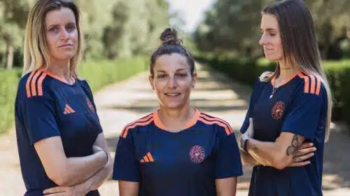 Three women in matching navy blue sports jerseys with orange accents stand on a sunlit, tree-lined path, arms crossed and facing the camera.