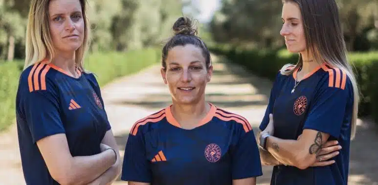 Three women in matching navy blue sports jerseys with orange accents stand on a sunlit, tree-lined path, arms crossed and facing the camera.