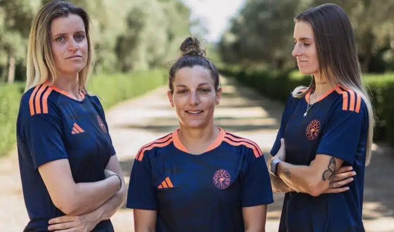 Three women in matching navy blue sports jerseys with orange accents stand on a sunlit, tree-lined path, arms crossed and facing the camera.