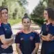Three women in matching navy blue sports jerseys with orange accents stand on a sunlit, tree-lined path, arms crossed and facing the camera.