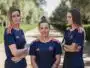 Three women in matching navy blue sports jerseys with orange accents stand on a sunlit, tree-lined path, arms crossed and facing the camera.
