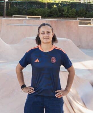 Screenshot | AllezPaillade.com Person standing with hands on hips in a skate park bowl, wearing a dark blue jersey with orange accents.